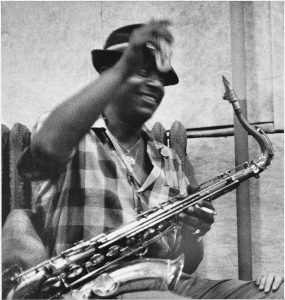 B&W photo, man seated, raising something blurry with hand, holding sax in the other/across his lap